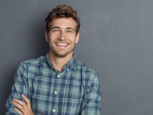 Young handsome man leaning against grey wall with arms crossed. Cheerful man laughing and looking at camera with a big grin. Portrait of a happy young man standing with crossed arms over grey background.
